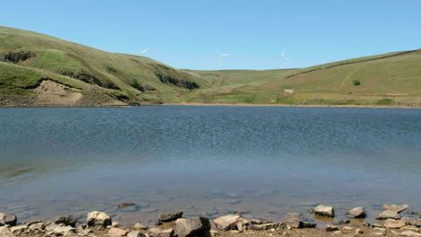 Wind-turbines-spin-in-the-background-behind-Magnificent-Green-hills-and-Beautiful-Calm-Lake,-Gorpley-Clough-Woods-Nature-Reserve