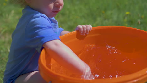 Toddler-joyfully-plays-and-splashes-water-from-a-bucket-on-a-hot,-bright-summer-day