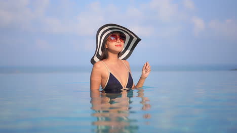 Hübsche-Asiatische-Frau-Mit-Hut-Und-Sonnenbrille-Im-Infinity-Pool,-Die-Sich-Langsam-Der-Kamera-Zuwendet,-Tourismuskonzept-In-Zeitlupe