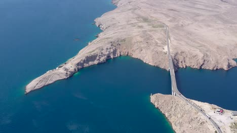 The-Paski-Most-Arch-bridge-linking-Pag-island-to-the-Croatian-mainland-with-Ruine-Fortica-abandoned-Fort-left,-Aerial-hovering-shot