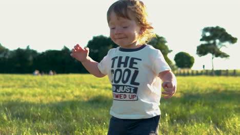 Slow-motion-of-adorable-blonde-toddler-walking-on-a-grass-field-in-warm,-soft-back-light
