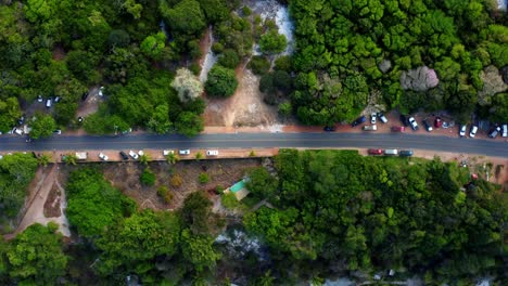 Bird's-eye-top-aerial-drone-view-of-a-small-road-surrounded-by-topical-trees-and-parked-cars-with-the-beach-of-Praia-do-Madeiro-near-Pipa-with-tourists,-umbrellas-and-golden-sand-in-Northern-Brazil