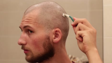 Young-bald-man-shaving-his-own-head-looking-in-the-mirror-with-disposable-razor,-still-medium-shot