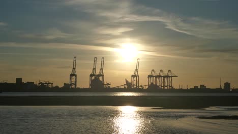 Shimmering-sunset-golden-river-water-with-British-harbour-port-cranes-silhouettes-skyline-wide-shot