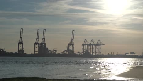 Tide-slow-moving-on-shimmering-sunset-golden-river-water-with-British-harbour-port-cranes-silhouettes-skyline