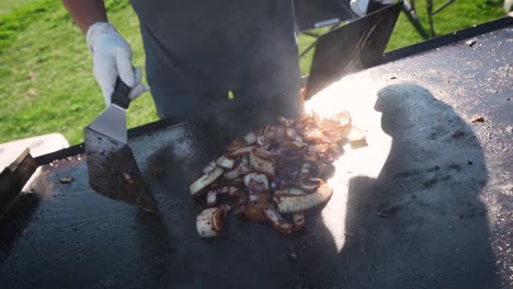Man-tossing-grilled-onions-with-spatulas-on-hot-grill-in-slow-motion,-close-up,-pull-away-for-backyard-bbq-with-friends