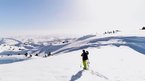One-person-standing-on-white-snow-looking-at-mountain-wearing-helmet,-holding-skis,-in-colorful-yellow-winter-pants,-Akoura,-Lebanon,-circle-aerial