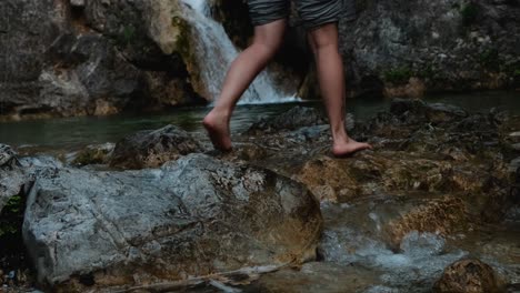 Young-woman-crosses-a-stream-barefoot-with-beautiful-waterfall-in-Mount-Olympus,-Greece