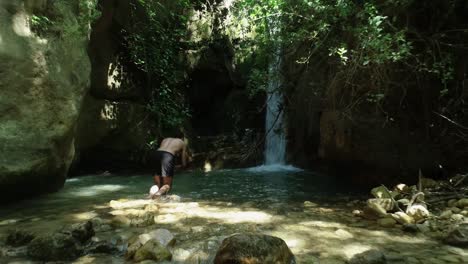 Young-Caucasian-shirtless-and-barefoot-man-wearing-black-bathing-suit-walks-to-fresh-mountain-pool-by-waterfall-and-dives-in,-low-backward-aerial