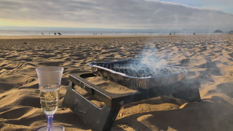 Smoke-sizzles-from-bbq-grill-on-beach-near-wine-glass,-low-angle