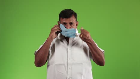 Close-up-indian-adult-man-wearing-medical-face-mask,-looking-to-camera-on-green-background