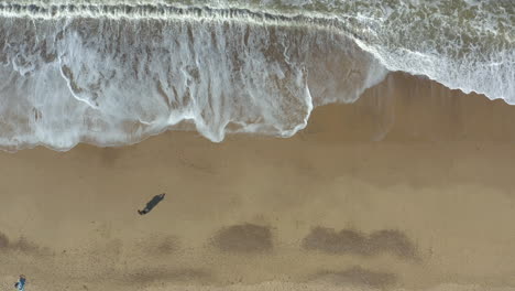 Aerial-shot-of-couple-with-elongated-shadows-standing-on-a-golden-sand-beach-and-admiring-the-sea
