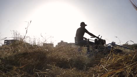 Low-angle-view-of-adult-male-farmer-riding-industrial-wheat-harvesting-machine-into-sunset-with-bright-white-sun-in-sky,-slow-motion-static