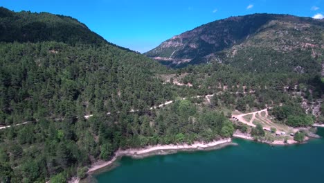 Tilt-down-aerial-reveal-shot-of-lake-Tsivlou-shore-surrounded-by-mountains-with-green-pine-trees