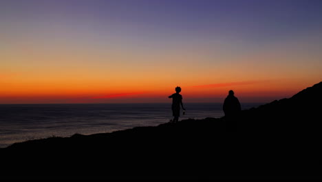 Silhouettes-of-adult-walk-along-shoreline-at-sunset,-wide