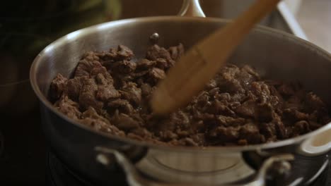 Cooking-brown-meat-pieces-in-silver-pot-on-kitchen-stove-with-wooden-slatted-spoon,-close-up-static