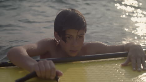 Junger-Mann-Umklammert-Das-Paddleboard,-Während-Er-Mit-Nachdenklichem-Blick-Im-See-Schwimmt