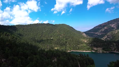 Aerial-shot-approaching-lake-Tsivlou-in-Peloponnese-mountains-of-Greece-with-a-bright-overcast-sky-and-clouds-casting-a-dark-shadow-on-pine-trees