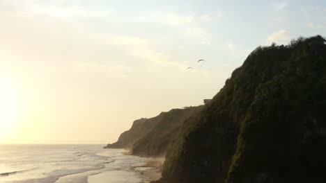 Two-paragliders-soaring-over-epic-cliffs-bordering-Melasti-beach,-South-Bali,-aerial-track