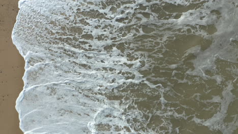Aerial-shot-of-big,-powerful,-high,-white-foam-waves-crashing-on-a-golden-sand-beach