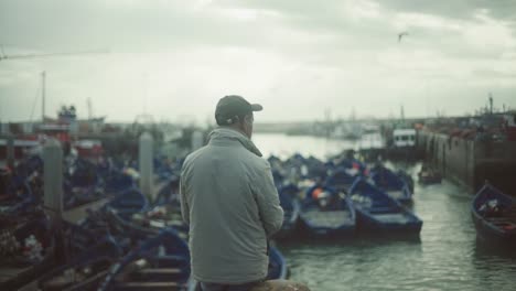 Mann-Steht-Und-Beobachtet-Vor-Dem-Hafen-Von-Essaouira-Marokko-Und-Sieht-Viele-Blaue-Fischerboote,-Authentisches-Afrikanisches-Meeresleben