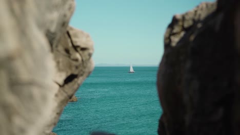 Peaceful-ocean-shore-view-with-sailboat-passing-by-through-rock-cliff-under-blue-sky-at-Cascais-Portugal-4K-steady-shot