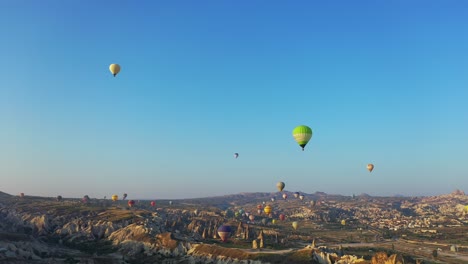 Hot-air-balloons-floating-above-Cappadocia-landscape-at-sunrise,-aerial-panning-left