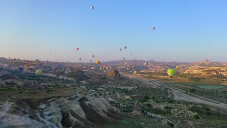 Many-hot-air-balloons-floating-above-Cappadocia-landscape-at-sunrise,-forward-aerial