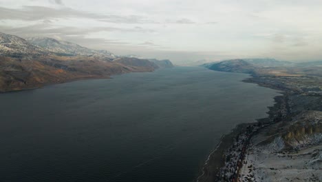 A-moody-wide-angle-shot-of-Kamloops-Lake-in-the-winter-surrounded-by-hilly-desert-landscape,-partially-covered-with-snow-and-a-train-travelling-along-it's-beaches