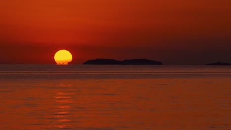 The-sun-is-setting-over-the-horizon-at-a-romantic-beach-at-the-Croatian-Mediterranean-seaside-as-a-big,-orange-ball-with-sailing-ships-passing-by