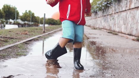 Little-boy-wearing-rubber-boots-splashing-water-from-a-puddle-to-the-camera-in-slow-motion