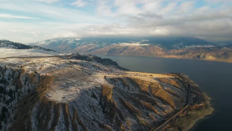 As-the-sun-begins-to-set-over-Kamloops-Lake-in-the-winter,-golden-hour-light-illuminates-the-partially-snow-covered-hilly-desert-terrain-around-the-lake