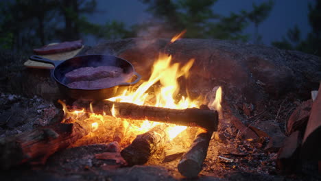 Outdoor-cooking-over-a-campfire,-grilling-steak-in-cast-iron-skillet-at-night