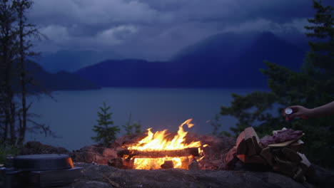Campfire-burns-at-night-in-mountain-campsite,-hand-seasons-food-for-cooking