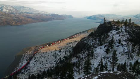 Cinematic-Shot-of-a-colorful-train-passing-by-Kamloops-Lake-in-the-winter,-snow-covered-rocky-cliffs-and-spruce-trees-are-visible-in-the-foreground