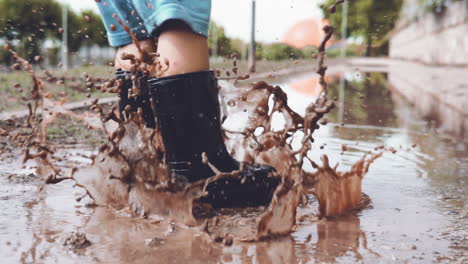 Close-up-video-of-kid-feet-wearing-rubber-boots-jumping-in-a-muddy-puddle-on-a-rainy-day-at-the-park