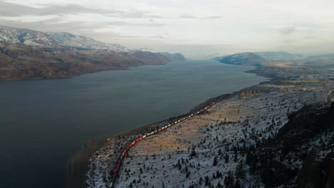 On-a-sunny-winter's-day,-a-colorful-train-drives-along-the-shores-of-Kamloops-Lake's-hilly-desert-landscape,-partially-covered-with-snow