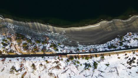Slow-Motion-Overhead-Shot-of-a-red-train-passing-by-the-beaches-of-Kamloops-Lake-during-golden-hour-throwing-long-shades,-amazing-scene-of-the-snow-covered-desert-environment