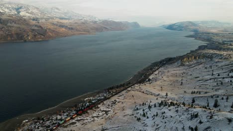 Stunning-capture-of-a-colorful-train-passing-along-the-shore-line-of-Kamloops-Lake-surrounded-by-a-hilly-desert-landscape-partially-covered-in-snow-during-the-early-winter-months-in-a-warm-sunlight
