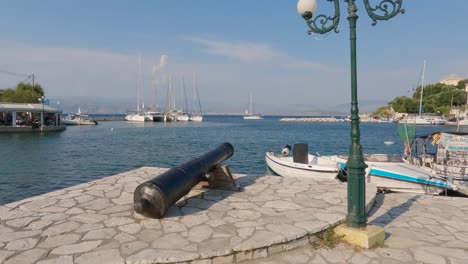 War-cannon-at-Picturesque-Kassiopi-promenade,-fishing-boats-moored-during-summer,-Corfu-island