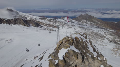 Aerial-flight-along-Austrian-flag-on-mountain-peak-and-lift-in-the-snowy-valley-of-Alps