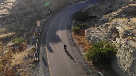 Luftaufnahme-Eines-Motorrads,-Das-Auf-Einer-Leeren,-Kurvenreichen-Autobahnstraße-Durch-Den-Hingol-Nationalpark-In-Der-Wüstenlandschaft-Von-Balochistan-Mit-Linseneffekt-Fährt