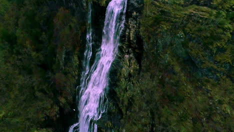 Aerial-drone-backward-moving-shot-over-Norwegian-waterfall-along-mountain-slope-covered-with-green-vegetation-at-daytime