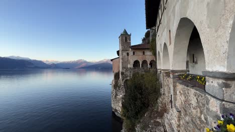 Panning-view-of-Santa-Caterina-Del-Sasso-hermitage-in-Italy-perched-on-Lake-Maggiore-with-snowy-Alps-mountains-range-in-background