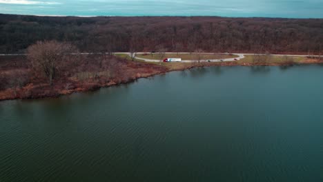 cinematic-aerial-of-a-red-semi-truck-and-white-trailer-parked-on-side-of-a-lake