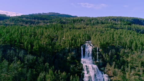 Aerial-backward-moving-shot-of-waterfall-from-top-of-a-mountain-surrounded-by-dense-green-forest-along-mountain-slope-at-daytime
