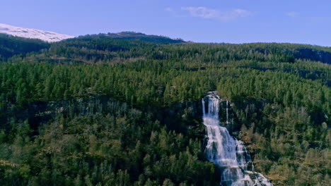 Aerial-drone-backward-moving-shot-of-a-waterfall-from-high-mountain-cliff-along-the-slope-on-a-summer-day