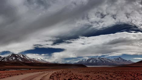 Wide-panoramic-scene-of-Bolivian-Andean-landscape-and-dirt-road-with-cloudy-sky-and-snowcapped-mountains-in-background,-Bolivia