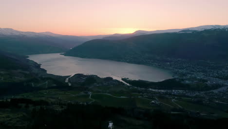 High-angle-shot-over-beautiful-norwegian-fjord-with-the-view-of-a-town-in-distance-with-sunset-in-the-background-during-evening-time