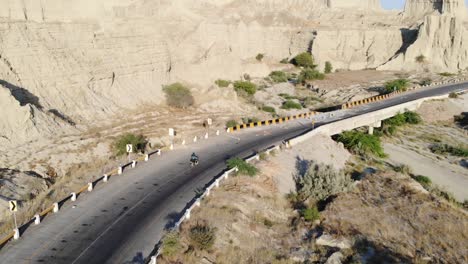 Tiro-De-Seguimiento-Aéreo-Detrás-De-La-Conducción-De-Motocicletas-A-Lo-Largo-De-Una-Carretera-Curva-Vacía-A-Través-Del-Parque-Nacional-Hingol-En-El-Paisaje-Desértico-De-Baluchistán